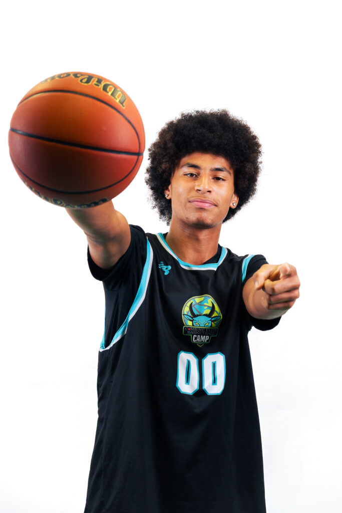 Jeune homme en maillot noir à détails bleus portant le logo « Combine Elite Basketball Camp » (numéro 00), tenant un ballon de basket Spalding et pointant vers l'avant, fond blanc.